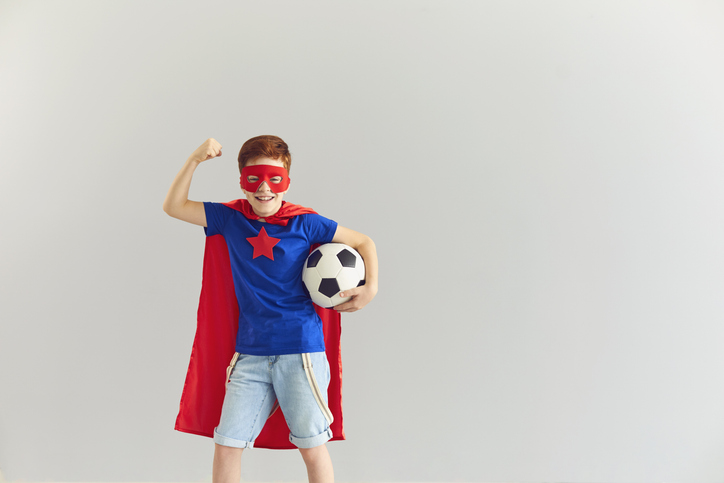 Smiling Boy In Superhero Costume Showing Power And Holding Soccer Ball In Hand, Copy Space